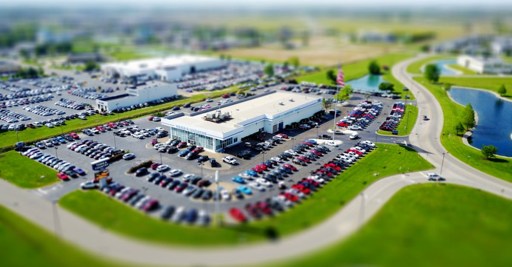 Aerial high-angle view of a bustling car dealership surrounded by parked cars in a green landscape.